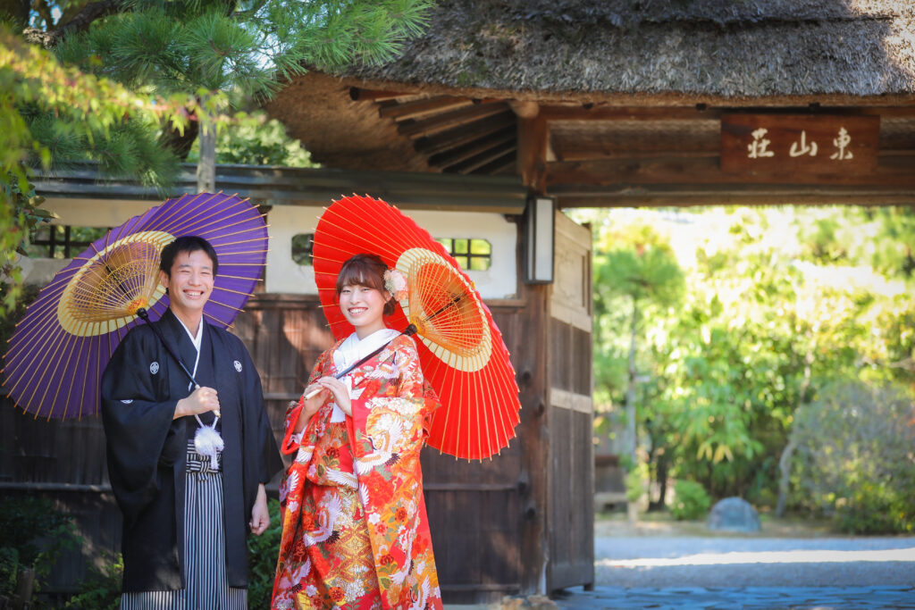 東山荘の門の前で、和傘をさしこちらを見て微笑む紋付き袴を着た新郎と色打掛を着た新婦