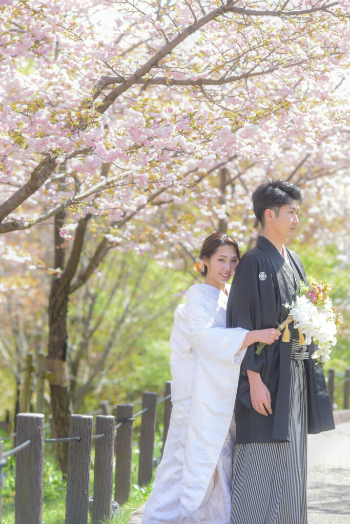 東山植物園内の桜の木の下で紋付き袴姿の新郎にハグをする白無垢姿の新婦