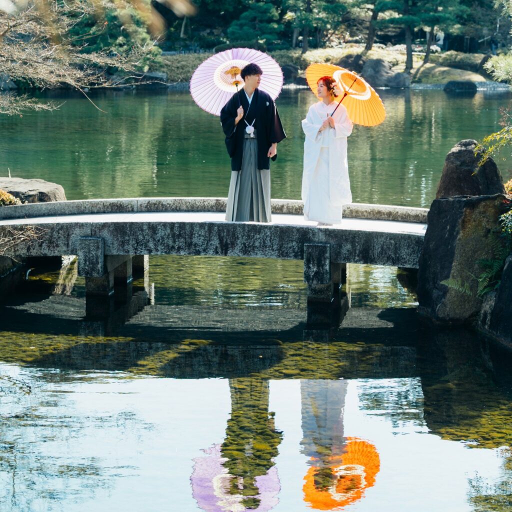 橋の上で和傘をさしながらお互いの顔を見つめる紋付き袴姿の新郎と白無垢姿の新婦