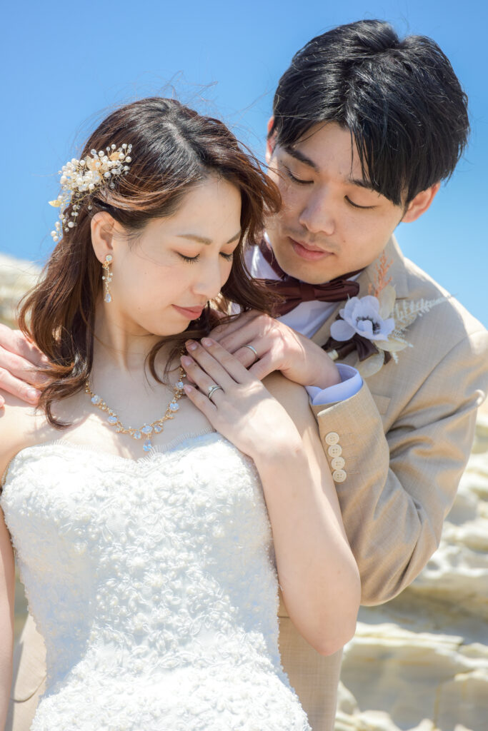 野間灯台周辺の岩場で顔を寄せ結婚指輪をしている手を重ねるタキシード姿の新郎とウェディングドレス姿の新婦